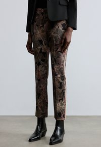 Pantaloni con motivo floreale neri con contorni rosa, vestibilità affusolata, texture liscia, indossati con stivaletti neri e giacca nera.