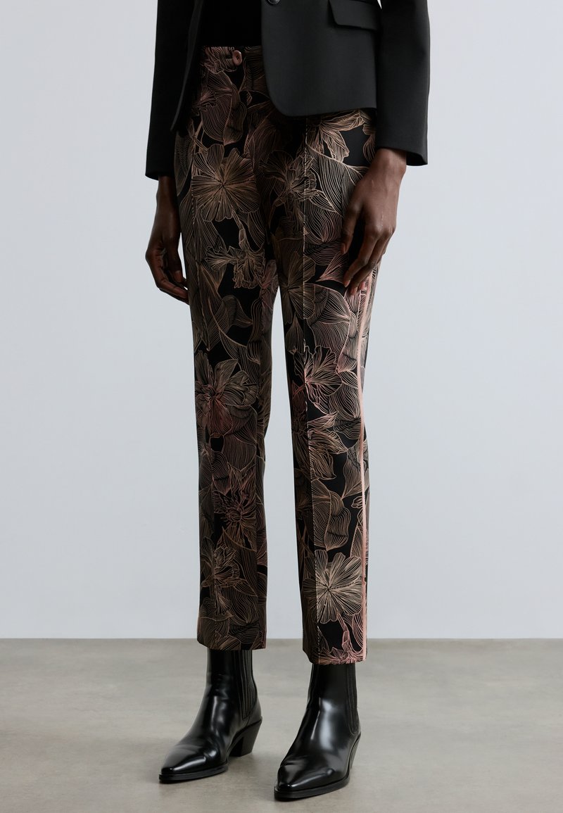 Pantaloni con motivo floreale neri con contorni rosa, vestibilità affusolata, texture liscia, indossati con stivaletti neri e giacca nera.