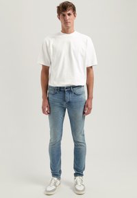 Una camiseta blanca de manga corta, jeans ajustados de color azul claro y zapatillas grises con detalles en blanco, de pie contra un fondo neutro.