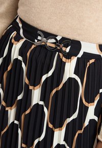 Faltrock mit einem abstrakten Muster in Schwarz, Weiß und Beige, versehen mit einer glänzenden silbernen runden Schnalle am Bund. Weicher Stofftextur.