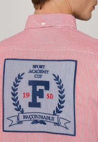 Rot und weiß gestreiftes Hemd mit einem blauen gestickten Patch, der Lorbeerblätter und den Buchstaben "F" sowie "SPORT ACADEMY CUP 1950" zeigt.