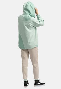 Licht muntgroene, lichte jas met een capuchon, voorzien van een textuurstof, gedeeltelijk zijpaneel en gele stikselaccenten.