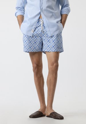 Hombre con camisa azul claro, pantalones cortos de baño azules con estampado y cordón, y zapatos marrones tejidos tipo slip-on, de pie con las manos en los bolsillos.