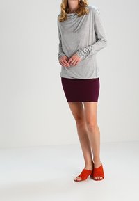 Maglia grigia a maniche lunghe con scollatura drappeggiata, abbinata a una gonna fitted di un intenso bordeaux scuro. Sandali a infradito in camoscio arancione. Texture morbide, design casual.