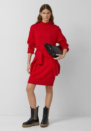 Rotes Strickkleid mit einem hohen Kragen, tailliert mit einem Gürtel und gerippten Texturen, kombiniert mit schwarzen Stiefeletten mit dicken Sohlen und Seiteneinsätzen.