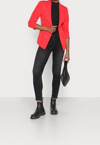 Blazer rojo con cuello estilo chal, dos bolsillos con cremallera, combinado con vaqueros pitillo negros y botines negros. Sosteniendo un bolso negro acolchado.