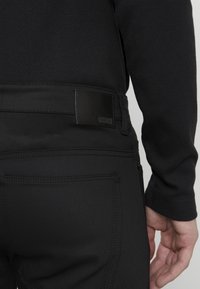 Černé kalhoty vyrobené z hladkého materiálu, s koženou značkou na pase, podrobným prošíváním a slim fit designem.