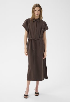 Žena v hnedom midi šatách s krátkymi rukávmi, zapínaných na gombíky vpredu, s opaskom v páse a čiernymi sandálmi s otvorenou špičkou, stojaca na bielom pozadí.