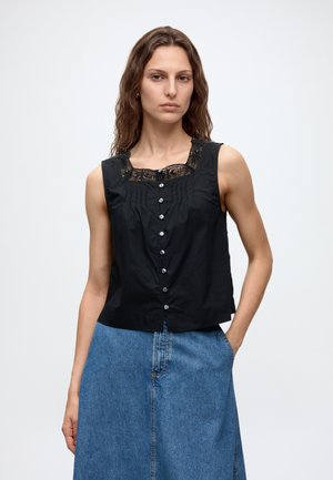 Kvinde med bølget hår iført en ærmeløs sort knapdet top med blonde detaljer og en høj taljet blå denimbuks, stående med den ene hånd i lommen.