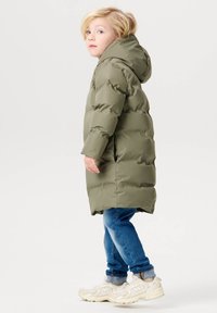 Olivgrüner Puffer-Mantel mit Kapuze, mit einem gesteppten Design und seitlichen Taschen, kombiniert mit blauen Jeans und hellen Sneakers.
