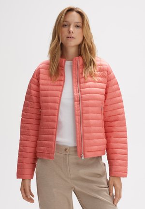 Gevoerde koraalkleurige jas met een staande kraag, rits aan de voorkant en ribbelstructuur. Gecombineerd met een witte top en beige broek.