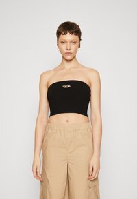 Černý žebrovaný strapless crop top s kovovým logem umístěným uprostřed. Modelka zkombinovala s béžovými cargo kalhotami. Jednoduchý, přiléhavý design.