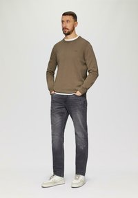 Bruine sweater, witte binnenlaag, grijze versleten jeans en witte sneakers. Model staat met de handen in de zakken tegen een effen achtergrond.