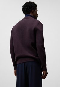 Sweter dzianinowy w głębokim purpurze, z wysokim kołnierzem, ściągaczami przy mankietach i u dołu, zestawiony z ciemnogranatowymi spodniami dla uzyskania warstwowego wyglądu.