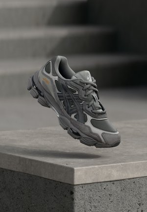Baskets Asics grises et noires en l'air au-dessus d'un rebord en béton avec des escaliers flous en arrière-plan.