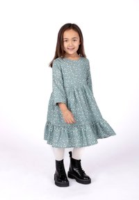 Helles türkisfarbenes Kleid mit weißem Blumenmuster, langem Ärmel und Rüschensaum. Kombiniert mit knielangen schwarzem Schnürstiefeln und cremefarbenen Strumpfhosen.