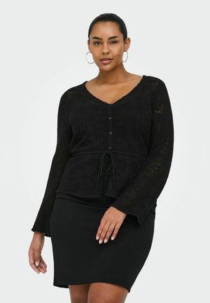 Nő fekete, horgolt gombos kardigánt visel, melyet derekán megkötött, továbbá fekete, testhez álló szoknyát, miközben világos, egyszínű háttér előtt pózol.