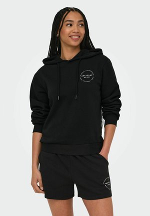 Donna con i capelli lunghi intrecciati che indossa una felpa nera con cappuccio e pantaloncini abbinati con logo circolare bianco sul petto e sulla coscia, sorridente e guardando verso destra.