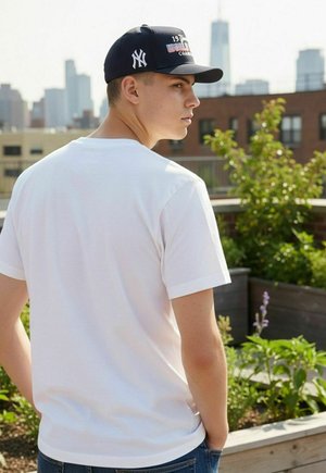 Jeune homme portant un t-shirt blanc et une casquette New York Yankees sombre, debout dans un jardin sur le toit avec la silhouette de la ville en arrière-plan.