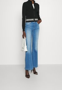 Giacca cropped nera con zip e fascia con logo, jeans a gamba larga blu chiaro, sandali neri con tacco e borsa bianca dalla forma strutturata.