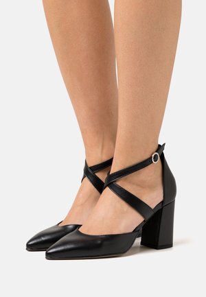 Chaussures à talons carrés en cuir noir avec bouts pointus et brides croisées à la cheville. Texture lisse et design minimaliste.