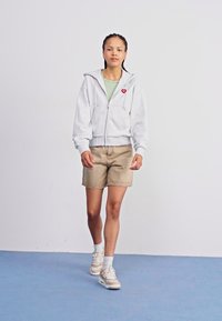 Graue Zip-Hoodie mit rotem Herz-Logo, grünem T-Shirt darunter, beigen Shorts und hellen Turnschuhen. Modell steht vor einem schlichten Hintergrund.