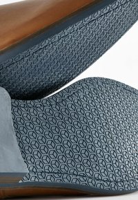 Zapato de cuero marrón con una suela de goma azul texturizada que presenta un patrón hexagonal. El diseño es elegante, con bordes suaves y costuras detalladas.