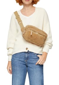 Tasche aus braunem Kunstfell in Crossbody-Form mit einem Reißverschlussfach, verstellbarem Riemen und weicher Textur. Getragen über einem weißen Pullover und Jeans.