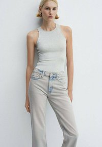 Tânără cu părul scurt blond, îmbrăcată într-un top fără mâneci de culoare gri deschis și blugi gri deschis cu talie înaltă, pe un fundal alb simplu.