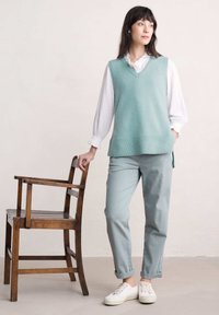 Un gilet en maille bleu clair sur une blouse blanche à volants, associé à un pantalon bleu clair et des baskets blanches, à côté d'une chaise en bois.