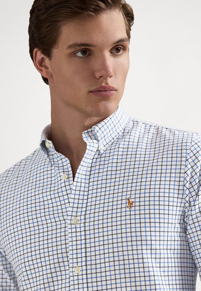 Λευκό πουκάμισο με κουμπιά, διακοσμημένο με μπλε καρό μοτίβο, γιακά με κουμπιά και πορτοκαλί κεντητό λογότυπο στο στήθος. Απαλό βαμβακερό ύφασμα.