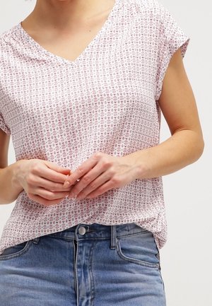 Kurzärmeliges V-Ausschnitt-Oberteil in Hellrosa mit detailreichen geometrischen Mustern. Weicher Stoff; lockere Passform, kombiniert mit blauen Jeans aus Denim.