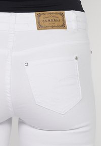 Jean en denim blanc avec une étiquette portant "KOROSHI" et des coutures décoratives sur les poches arrière, qui possèdent des accents en métal argenté.