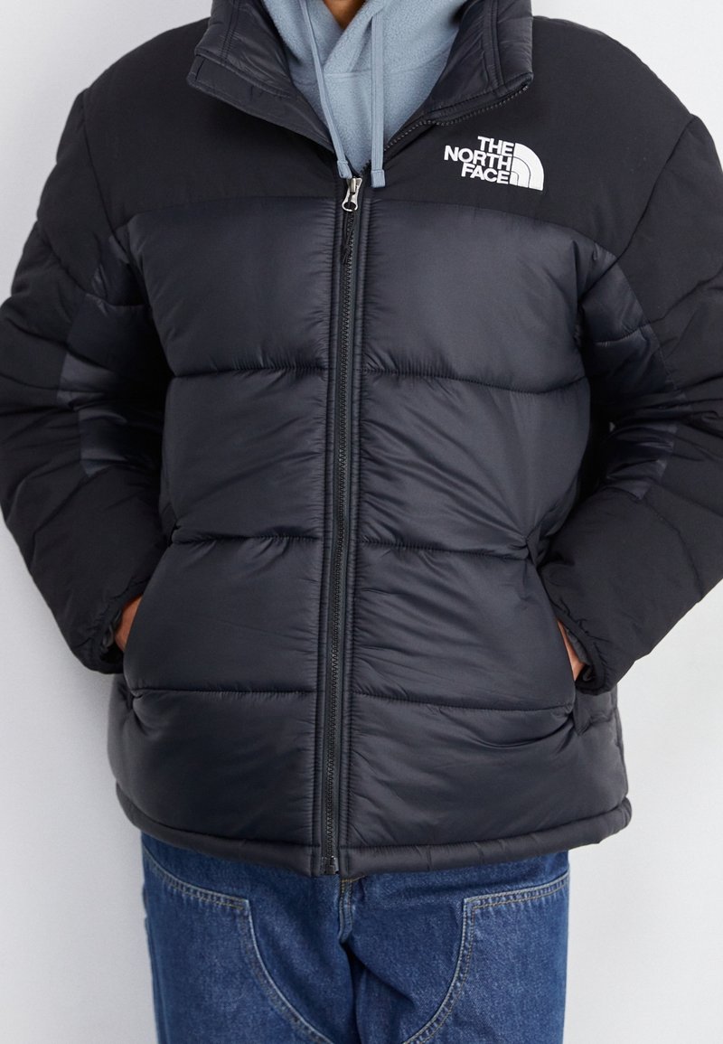 Mężczyzna w czarnej kurtce puchowej z logo The North Face, nosi niebieską bluzę pod spodem i niebieskie dżinsy, ręce schowane w kieszeniach kurtki.