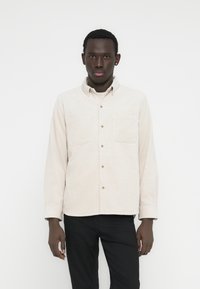 Világosbézs gombos ing két mellzsebbel, texturált anyagból és gallérral. Fekete nadrággal párosítva. Egyszerű, casual kialakítás.