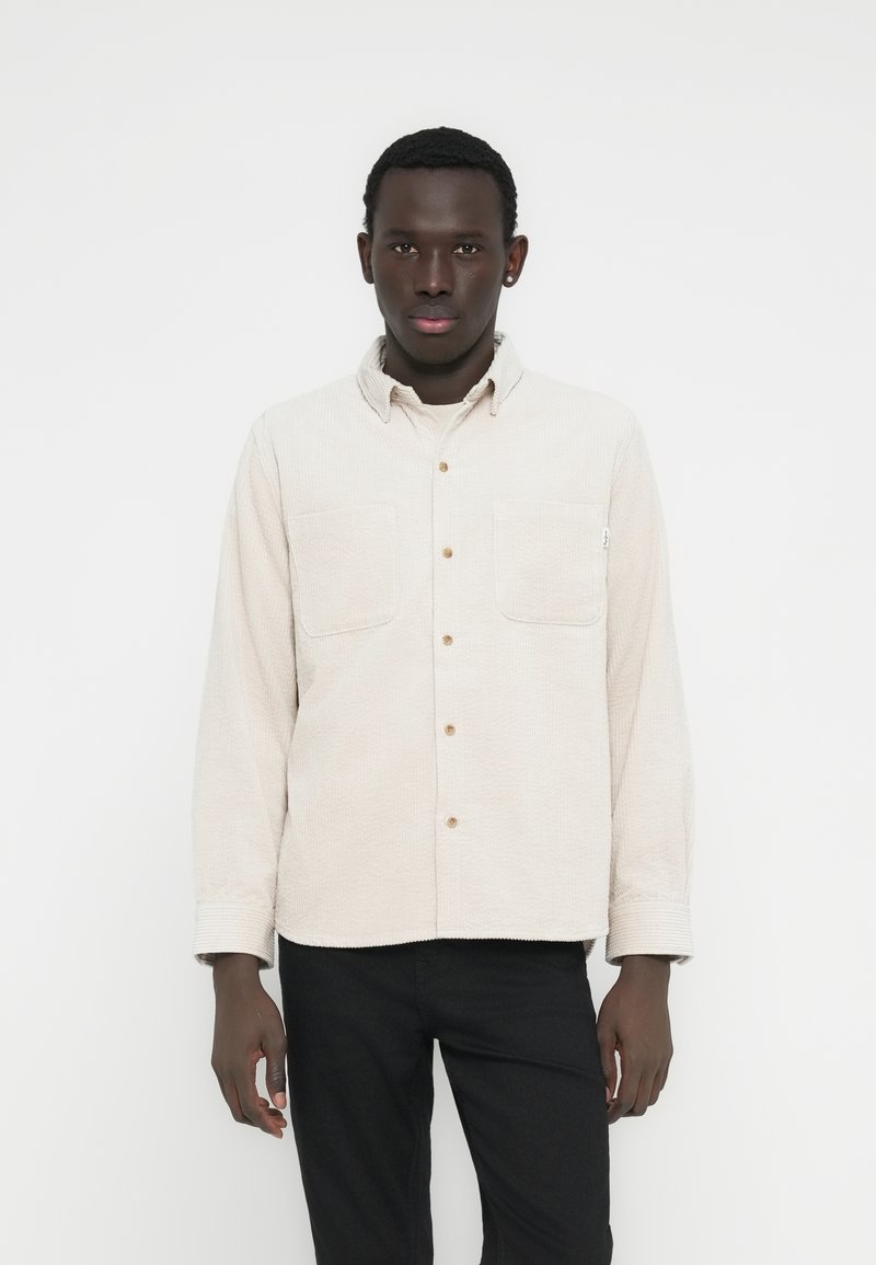 Világosbézs gombos ing két mellzsebbel, texturált anyagból és gallérral. Fekete nadrággal párosítva. Egyszerű, casual kialakítás.