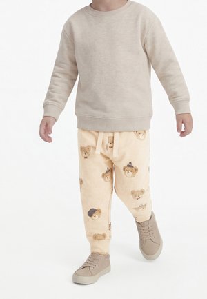 Barn iført beige sweatshirt og lysegule bukser med bamseansigter, går i taupe sneakers mod en hvid baggrund.