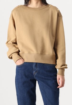 Nainen pukeutuneena kamelinväriseen collegepaitaan, jossa on kullanväriset napit hartioilla, ja sinisiin farkkuihin, käsi taskussa, yksiväristä taustaa vasten.