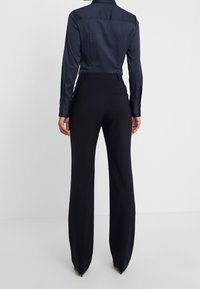 Chemise bleu marine à manches longues avec une texture lisse et brillante, agrémentée de poignets à boutons, associée à un pantalon noir à jambes larges avec une poche arrière.