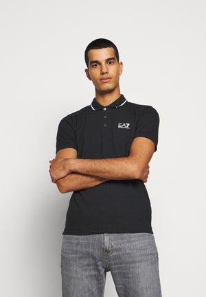 Camisa polo negra con ribete blanco en el cuello y logo de EA7 en el pecho, combinada con jeans gris claro. Textura de tela suave.
