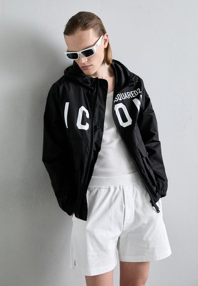 Mustat hupparitakki, jossa on suuret valkoiset kirjaimet "ICON" ja "DSQUARED2"; yhdistetty valkoisiin shortseihin ja valkoiseen hihattomaan paitaan, joissa on sileä pinta.
