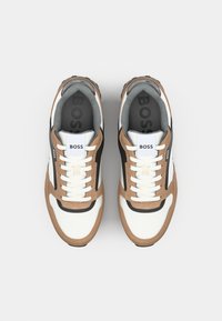 Witte en beige sneakers met zwarte accenten. Kenmerken zijn een gestructureerde bovenkant, witte veters en een logo op de tong en inlegzool.