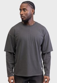 Tricou scurt cu mânecă peste dimensiune, de culoare gri închis, stratificat peste un strat de bază cu mânecă lungă, realizat din bumbac, având un etichetă pe marginea de jos.