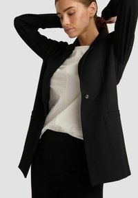 Schwarzer Blazer aus glattem Stoff mit taillierter Passform, Ein-Knopf-Verschluss und zwei Seitentaschen, getragen über einer weißen Bluse.