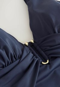 Marineblå badetop med rynket stof, med en guldfarvet lås og glat tekstur, designet til justerbar pasform og støtte.