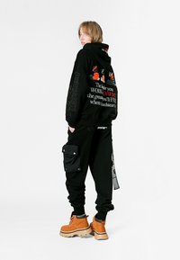 Joven con sudadera negra con texto y gráficos en rojo y blanco, pantalones cargo negros y botas gruesas marrones, de pie contra un fondo blanco.