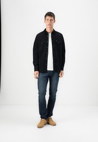 Camisa negra de botones con dos bolsillos en el pecho sobre una camiseta blanca, combinada con jeans azul oscuro y zapatillas color beige. Un look simple y casual.