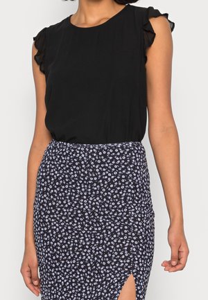 Eine Frau trägt eine schwarze ärmellose Bluse mit Rüschen an den Schultern und einen schwarzen Rock mit weißem Blumenmuster und einem seitlichen Schlitz.