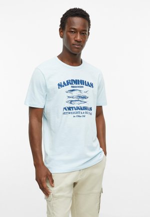 Jonge man draagt een lichtblauw T-shirt met een blauwe sardinesblik-afbeelding en beige cargobroek, staand tegen een witte achtergrond.