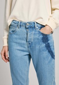 Jeans de denim azul claro com um padrão ligeiramente desgastado na frente. Design clássico de cinco bolsos, com costura visível e fecho com botão.